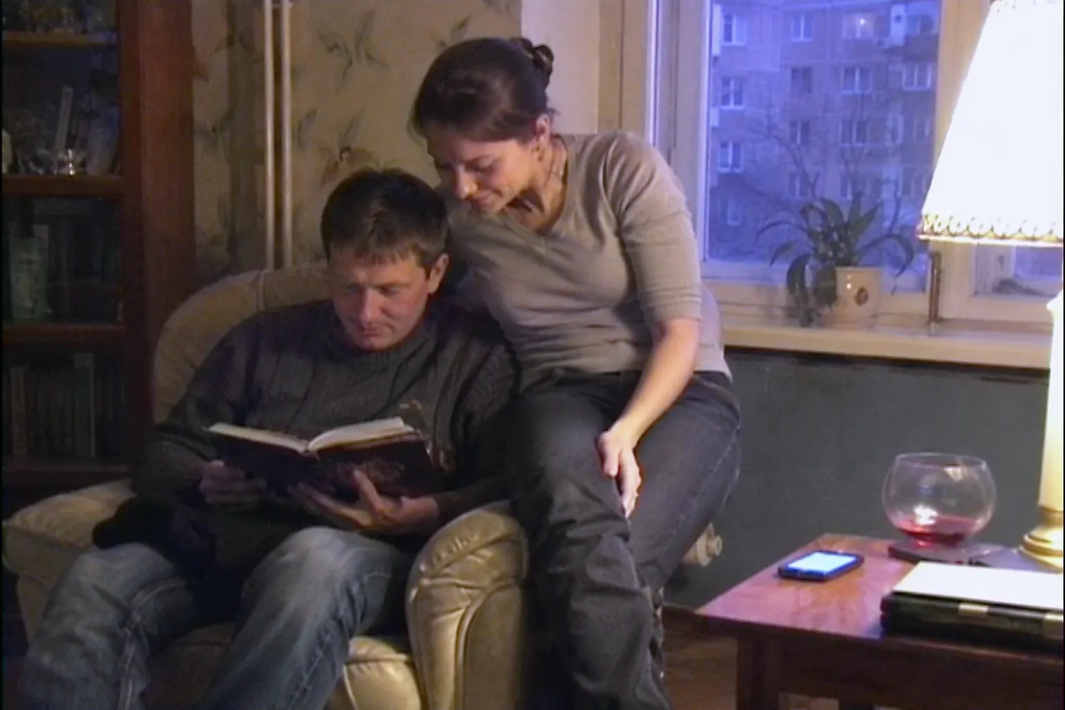 Девушка и мужчина вместе читают книгу в кресле у окна поздним вечером